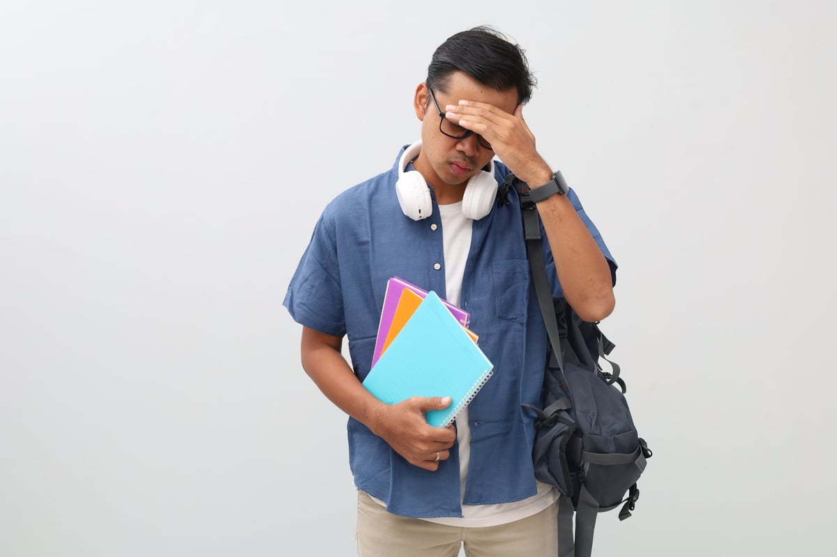 Young Asian male student looking frustrated and tired, standing isolated on white background