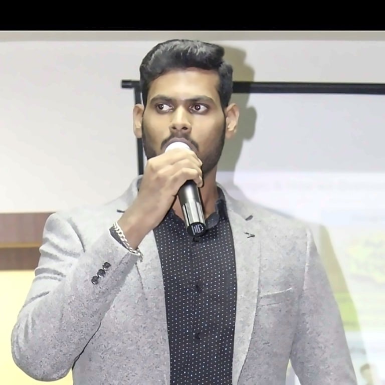 Man in light gray blazer holding microphone while speaking at an indoor event